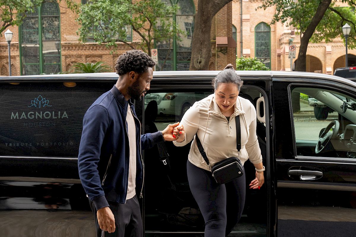 A person helps another out of a black van in front of a brick building with arched windows and trees lining the street.